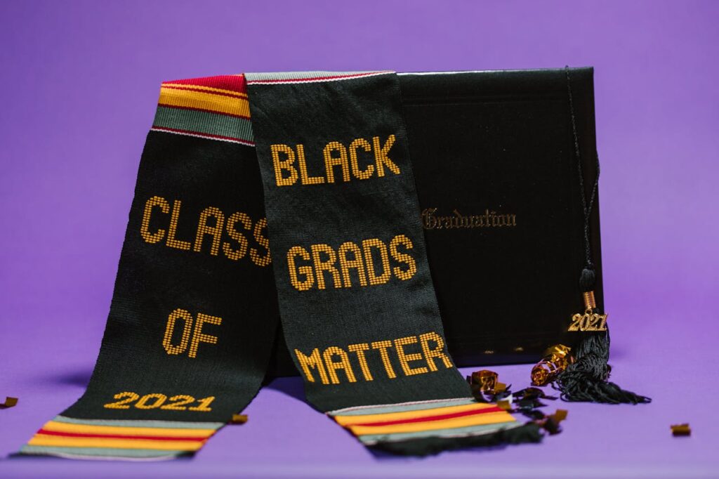 Graduation sash with 'Black Grads Matter' and cap on a purple backdrop, symbolizing achievement.